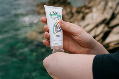 A person applies Nodol Aspirin-Free Pain Cream from a tube labeled 1936 Original onto their arm, seeking relief from joint pain. The background features the sea and rocky cliffs, evoking a coastal setting enhanced with natural ingredients.