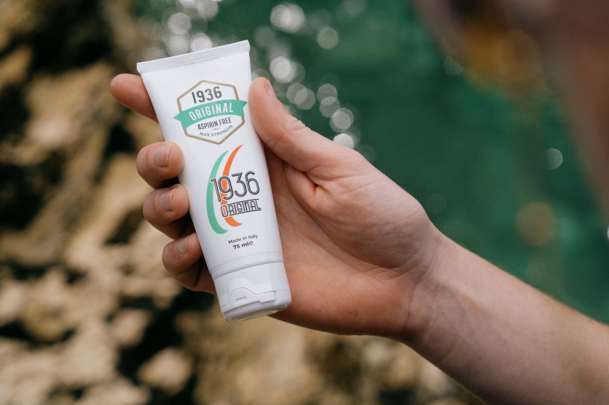 A hand holds a 75ml white tube labeled "1936 Original Nodol Aspirin-Free Pain Cream," boasting a natural formula. The packaging features green and orange accents, indicative of its muscle pain relief formulation. A blurred background of greenery and water complements the product's essence.